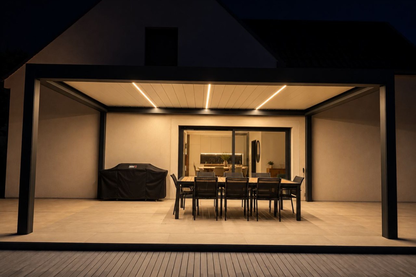 Terrasse couverte avec table à manger, barbecue et vue sur la cuisine intérieure, illuminée par des LED.