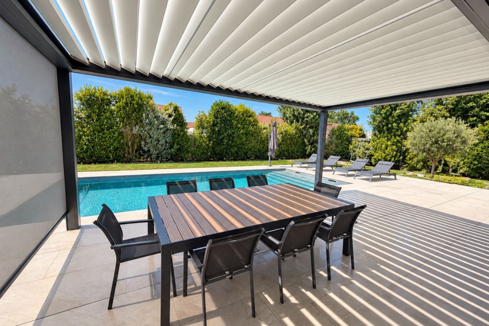 Terrasse couverte avec table et chaises, sous un auvent à lames verticales, face à une piscine entourée de verdure.
